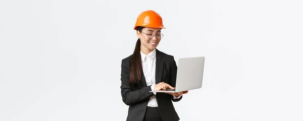 Sonriendo exitosa mujer asiática ingeniero industrial, gerente de fábrica en casco de seguridad y traje de negocios utilizando ordenador portátil, comprobando con el proyecto o planos en la pantalla — Foto de Stock