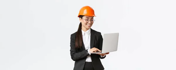 Lachen succesvolle vrouwelijke Aziatische industrieel ingenieur, fabrieksmanager in veiligheidshelm en business suit met behulp van laptop computer, controleren met project of blauwdrukken op het scherm — Stockfoto