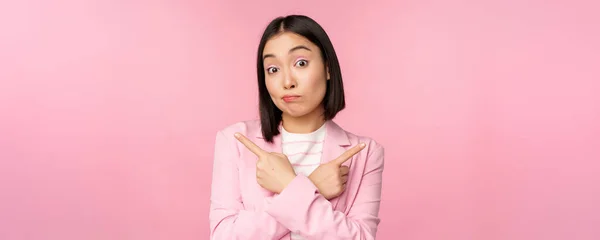 Conceito de decisão. Jovem mulher de negócios asiática, trabalhador corporativo apontando dedos lateralmente, duas direções, mostrando variantes, olhando indeciso ao escolher, fundo rosa — Fotografia de Stock