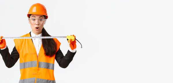 놀라고 흥분 한 동양 여성 건설 기술자, 안전모와 제복을 입은 산업 노동자, 구조를 측정하고 줄자를 잡고 카메라를 응시하며 백지 배경에 놀란 모습 — 스톡 사진