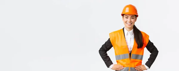 Exitosa joven mujer de negocios asiática, arquitecta en casco de seguridad, chaqueta reflectante, de pie confiada y decidida, sonriente asegurada, construyendo casas, posando sobre fondo blanco — Foto de Stock