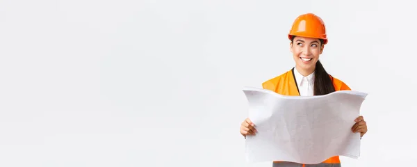 Creativa asiática mujer arquitecta imaginando su plan, de pie en casco de seguridad y mirando hacia otro lado reflexivo mientras lee planos, imaginando edificio después de las obras de construcción de acabado, fondo blanco — Foto de Stock