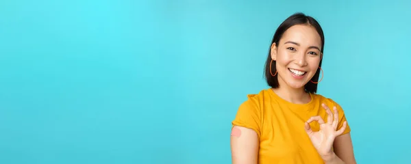 Vaccine campaign from covid-19. Happy healthy asian girl showing okay sign and smiling, bandaid on shoulder from coronavirus vaccination, blue background — Stock Photo, Image