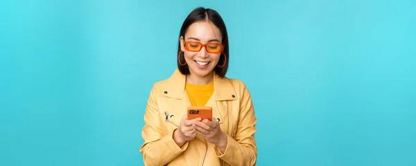 Smiling asian girl in sunglasses, using smartphone app, holding mobile phone, standing over blue background — Stock Photo, Image