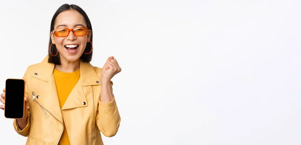 Felice ragazza asiatica in occhiali da sole, mostrando schermo del telefono cellulare, interfaccia smartphone, ridere e sorridere, festeggiare, in piedi su sfondo bianco — Foto Stock