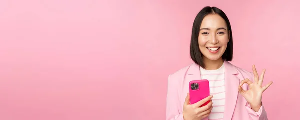 Sourire asiatique femme d'affaires montrant signe correct tout en utilisant l'application de téléphone mobile, recommander l'application smartphone, debout sur fond rose — Photo