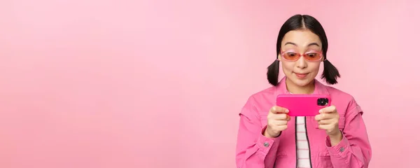 Портрет щасливої азіатки, що грає на смартфоні, дивиться відео на мобільний додаток, стоїть над рожевим тлом — стокове фото