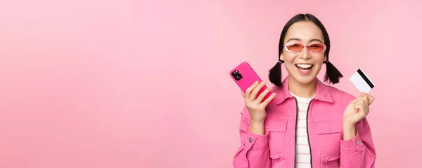 Сучасна красива азіатська дівчина, сміється і посміхається з мобільним телефоном, кредитною карткою, покупками в Інтернеті, платить зі смартфоном, стоїть на рожевому фоні — стокове фото