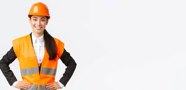 Exitosa joven mujer de negocios asiática, arquitecta en casco de seguridad, chaqueta reflectante, de pie confiada y decidida, sonriente asegurada, construyendo casas, posando sobre fondo blanco — Foto de Stock