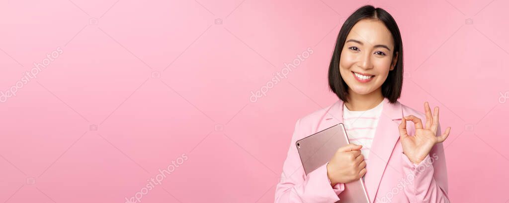 Portrait of corporate woman, girl in office in business suit, holding digital tablet, showing okay, recommending company, standing over pink background.