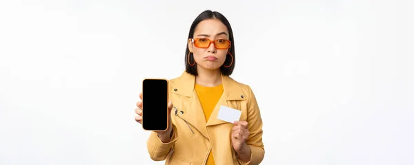 Triste chica asiática en gafas de sol, mostrando interfaz de aplicación de teléfono inteligente, tarjeta de crédito, mirando decepcionado, de pie sobre fondo blanco — Foto de Stock