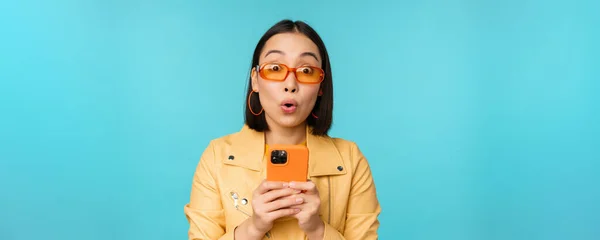 Obraz azjatyckiej dziewczyny w okularach przeciwsłonecznych, wyglądającej zdumiewająco i pod wrażeniem, nagrywającej wideo lub robiącej zdjęcia na smartfonie, stojącej nad niebieskim tłem — Zdjęcie stockowe