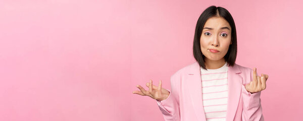 Portrait of young asian business woman, saleswoman shrugging shoulders and looking confused, clueless of smth, standing over pink background