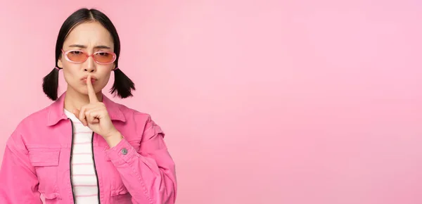Menina coreana moderna em roupa elegante primavera, óculos de sol, mostrando shush, sinal de silêncio, pressione o dedo para os lábios, gesto tabu, de pé sobre fundo rosa — Fotografia de Stock