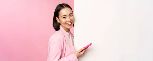 Afbeelding van Koreaanse vrouwelijke ondernemer in pak, staande in de buurt van info muur, reclame aan boord, holding smartphone en glimlachen, poseren over roze achtergrond — Stockfoto