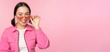 Göz giysisi reklamı. Modern Asyalı zarif bir kız güneş gözlüğüne dokunur, pembe giyer, stüdyo arka planında poz verir. Boşluğu kopyala