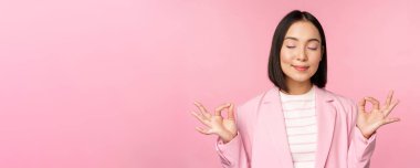 Sakin olun. Gülümseyen genç Asyalı kadın meditasyon yapıyor, yoga yapıyor, iş yerinde farkındalık ve gevşeme gösteriyor, nefes alıyor, keyifli bir şekilde havayı soluyor, pembe arka planda duruyor.