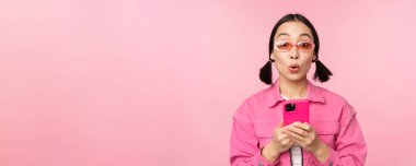 Moda güneş gözlüklü Koreli bir kızın portresi cep telefonu kullanıyor, akıllı telefon uygulaması kullanıyor, pembe arka planda duruyor.