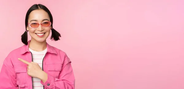 Retrato de sorrindo ásia menina no elegante roupa, óculos de sol, apontando dedo esquerdo, mostrando anúncio, banner, pé sobre rosa fundo — Fotografia de Stock