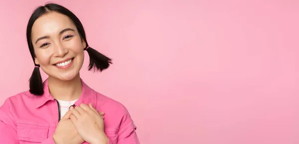 Menina coreana bonito com pães de cabelo bobo, olhando feliz e grato, obrigado pose, de mãos dadas no coração lisonjeado, posando contra fundo rosa — Fotografia de Stock