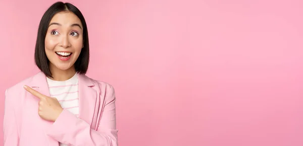 Entusiasta joven mujer de negocios asiática, empleado de oficina señalando con el dedo a la izquierda, mirando banner, anuncio con sonrisa feliz, mostrando publicidad, fondo rosa — Foto de Stock