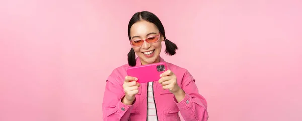 Retrato de la chica asiática feliz jugando en el teléfono inteligente, viendo videos en la aplicación del teléfono móvil, de pie sobre fondo rosa — Foto de Stock