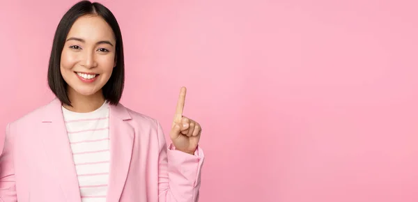 Retrato de mulher de negócios asiática apontando os dedos para cima e sorrindo, mostrando o logotipo da empresa de negócios, informações em cima, de pé sobre fundo rosa — Fotografia de Stock