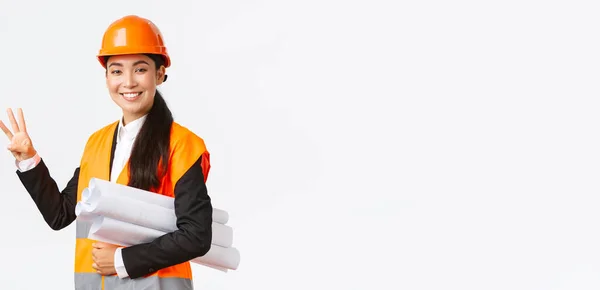 Χαμογελώντας Ασιάτισσα αρχιτέκτονα στο εργοτάξιο, χτίζοντας κτήματα, δείχνοντας το νούμερο τρία, μεταφέρουν σχέδια φορώντας κράνος ασφαλείας, δίνοντας σταθερό χρόνο για την εργασία που γίνεται, λευκό φόντο — Φωτογραφία Αρχείου