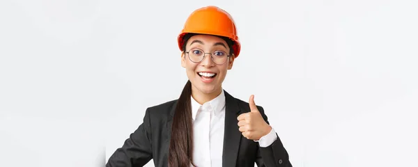 Close-up of professional pleased asian female chief engineer, architect in business suit and safety helmet showing thumbs-up in approval, give permission, recommend construction company — Stock Photo, Image