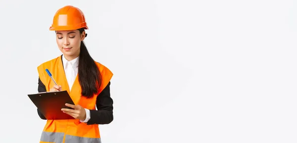 Agréable ingénieur de construction asiatique, architecte prenant des notes dans le presse-papiers, écrivant quelque chose pendant l'inspection à la zone du bâtiment, portant un casque de sécurité, femme d'affaires inspecter les travailleurs — Photo