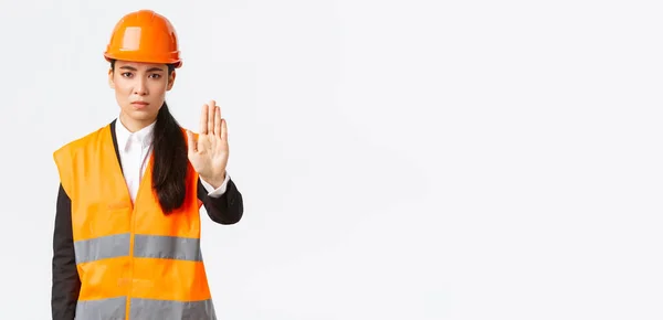 Ernstig uitziende Aziatische vrouwelijke architect, bouwmanager op het werk gebied dragen van een veiligheidshelm, tonen stop gebaar, verbieden van actie, verboden huisvredebreuk, witte achtergrond — Stockfoto