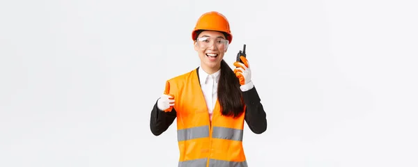 Soddisfatto felice sorridente asiatica ingegnere femminile, tecnico industriale in casco di sicurezza e uniforme mostrando pollici in su mentre loda grande lavoro utilizzando walkie-talkie, dare il permesso di lavorare — Foto Stock