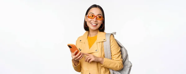 Snygg ung asiatisk kvinna turist, resenär med ryggsäck och smartphone ler mot kameran, poserar mot vit bakgrund — Stockfoto