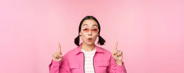 Närbild porträtt av asiatisk kvinna kippar, ser förvånad, pekar finger åt banner, reklam, står över rosa bakgrund — Stockfoto