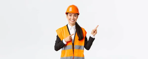 Construção, construção e conceito industrial. Sorrindo engenheiro asiático feminino em capacete de segurança e roupas reflexivas mostrando objeto, apontando dedos direito, vendedora introduzir propriedade para o cliente — Fotografia de Stock