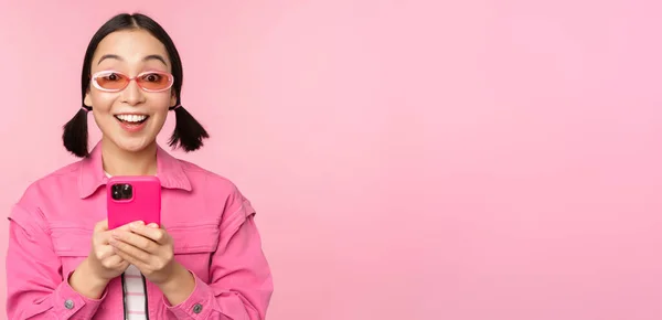 Ritratto di ragazza coreana con eleganti occhiali da sole che reggono il telefono cellulare, utilizzando l'app per smartphone, in piedi su sfondo rosa — Foto Stock