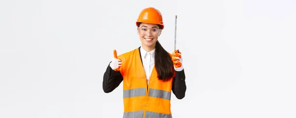 Sonriente mujer confiada asiática ingeniero de construcción, arquitecto en casco de seguridad, uniforme, mostrando cinta métrica y pulgares hacia arriba, satisfecho con buenos resultados, mantenerse bien hecho para el equipo, fondo blanco — Foto de Stock