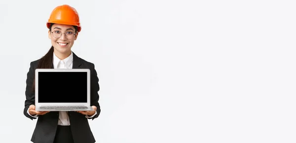 Sorrindo profissional engenheiro asiático feminino introduzir plano de construção para investidores ou clientes, de pé em capacete de segurança e terno mostrando tela do laptop com sorriso satisfeito, fundo branco — Fotografia de Stock