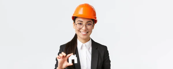 Közelkép boldog ázsiai nő engingeer, ingatlan ügynök sisak és üzleti ruha holding ház miniatűr és mosolygós, építész dolgozik felújítási projekt, fehér háttér — Stock Fotó