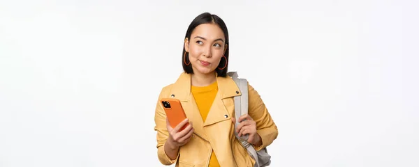 Asiático menina viajante com mochila, segurando telefone celular, usando o aplicativo do smartphone, olhando pensativo, de pé sobre fundo branco — Fotografia de Stock