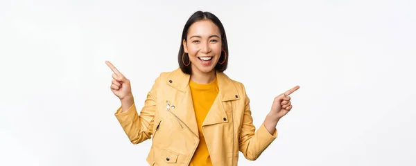 Feliz hermosa chica asiática señalando los dedos izquierda y derecha, mostrando el logotipo de la bandera, demostrando venta, de pie en chaqueta amarilla sobre fondo blanco —  Fotos de Stock