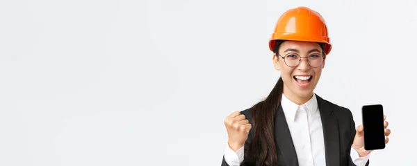 Bem sucedido vencedor asiático engenheiro feminino, arquiteto em capacete de segurança e terno, mostrando tela do smartphone, punho bomba e gritando sim de alegrar-se, triunfando sobre a realização, fundo branco — Fotografia de Stock