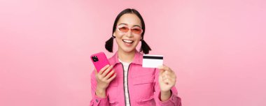 Modern, güzel Asyalı kız, cep telefonu, kredi kartı, online alışveriş, akıllı telefon ile ödeme, pembe arka planda durmak