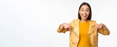 Mutlu gülümseyen Asyalı kızın portresi, parmakları aşağıda ve logoyu gösteriyor, pankartı gösteriyor, sarı ceketin içinde beyaz arka planda duruyor.