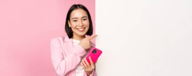 Takım elbiseli gülümseyen Asyalı şirket kadını, elinde akıllı telefon, tahtayı işaret eden, boş beyaz duvarda çizelge ya da bilgi logosunu gösteren, pembe arka planda duran