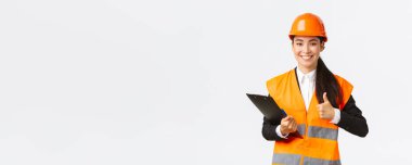 Tatmin olmuş Asyalı kadın inşaat mühendisi, teftiş sonuçlarından memnun. İnşaat alanında giysi ve kask giyiyor, notlar yazıyor ve onayını veriyor.