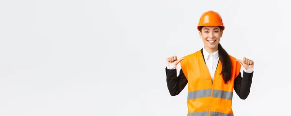 Successful proud smiling asian female construction manager, engineer in safety helmet pointing at herself, show-off. Architect recommend personal assistance, bragging accomplishments — Stock Photo, Image