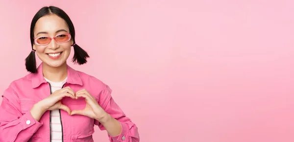 Gyönyörű koreai nő modell divatos napszemüveg, mutatja szív, ellátás jel, Szeretem gesztus, áll több mint rózsaszín háttér — Stock Fotó