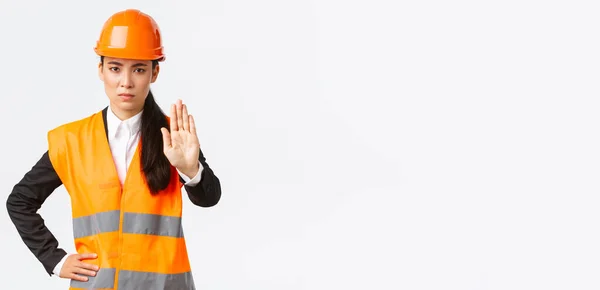 Ernstig uitziende Aziatische vrouwelijke architect, bouwmanager op het werk gebied dragen van een veiligheidshelm, tonen stop gebaar, verbieden van actie, verboden huisvredebreuk, witte achtergrond — Stockfoto
