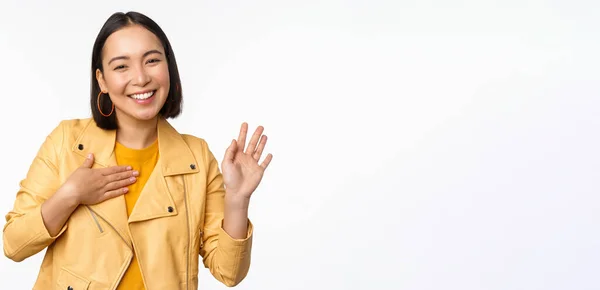 Bild av vänlig asiatisk flicka i snygg gul rock, höja armen, presentera sig, hälsning, viftande hand, säger hej, står över vit bakgrund — Stockfoto